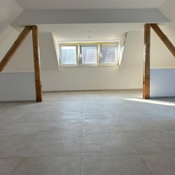 Firma ogólnobudowlana Artur Tchórzewki - Jasne poddasze z drewnianymi belkami i nową, szarą podłogą z płytek. Trzy okna dachowe wpuszczają światło słoneczne. Świeżo wykończone wnętrze.