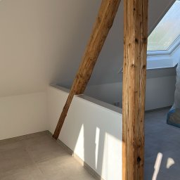 Firma ogólnobudowlana Artur Tchórzewki - Jasne poddasze po renowacji z drewnianą belką konstrukcyjną, szarą podłogą z płytek i oknem dachowym. Świeżo pomalowane ściany w bieli. Promienie słoneczne podkreślają fakturę drewna.