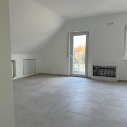 Firma ogólnobudowlana Artur Tchórzewki - Jasne wnętrze poddasza z szarymi płytkami podłogowymi i białymi ścianami. Widoczna skrzynka z instalacją grzewczą i okno balkonowe z widokiem na dom.