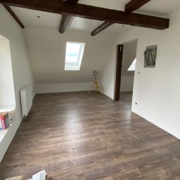 Firma ogólnobudowlana Artur Tchórzewki - Jasne poddasze po remoncie: drewnopodobne panele podłogowe, białe ściany, widoczne drewniane belki stropowe i okno dachowe. Częściowo widoczne materiały budowlane.