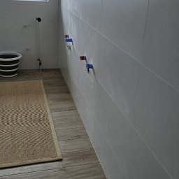 Firma ogólnobudowlana Artur Tchórzewki - Wykończona łazienka z szarymi płytkami na ścianach i podłodze imitującej drewno. Widoczne przyłącza wodne i okno z widokiem na zewnątrz.