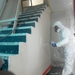 Firma ogólnobudowlana Artur Tchórzewki - Malowanie natryskowe ściany na klatce schodowej przez pracownika w kombinezonie ochronnym. Widoczna drabina i zabezpieczone schody.