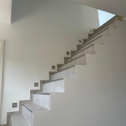 Firma ogólnobudowlana Artur Tchórzewki - Wykończone schody z szarymi płytkami i oświetleniem punktowym na ścianie. Minimalistyczny design, jasne wnętrze, nowoczesne wykończenie.