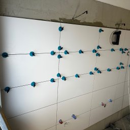 Firma ogólnobudowlana Artur Tchórzewki - Białe płytki ścienne w trakcie montażu, z użyciem systemu poziomowania płytek (niebieskie klipsy i kliny). Widoczne otwory na instalacje. Ściana częściowo wykończona.