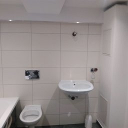 Firma ogólnobudowlana Artur Tchórzewki - Jasna łazienka po remoncie: białe płytki, umywalka, toaleta i wanna z hydromasażem. Widoczne niedokończone podłączenia elektryczne.