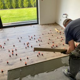 Firma ogólnobudowlana Artur Tchórzewki - Układanie paneli podłogowych z użyciem poziomicy i klipsów dystansowych w jasnym wnętrzu z dużym oknem na ogród. Widoczny fragment nałożonego kleju.