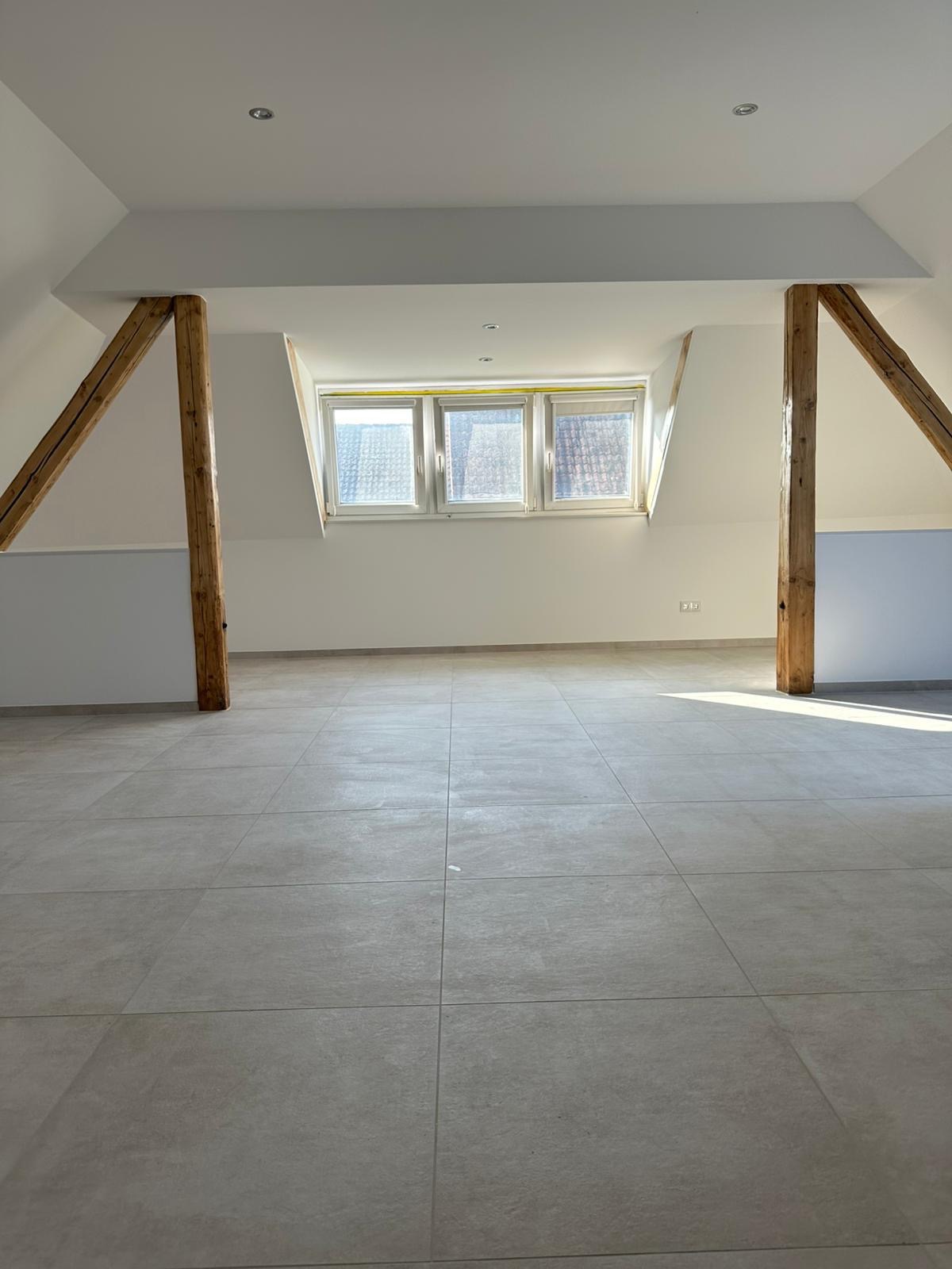 Jasne poddasze z drewnianymi belkami i nową, szarą podłogą z płytek. Trzy okna dachowe wpuszczają światło słoneczne. Świeżo wykończone wnętrze.