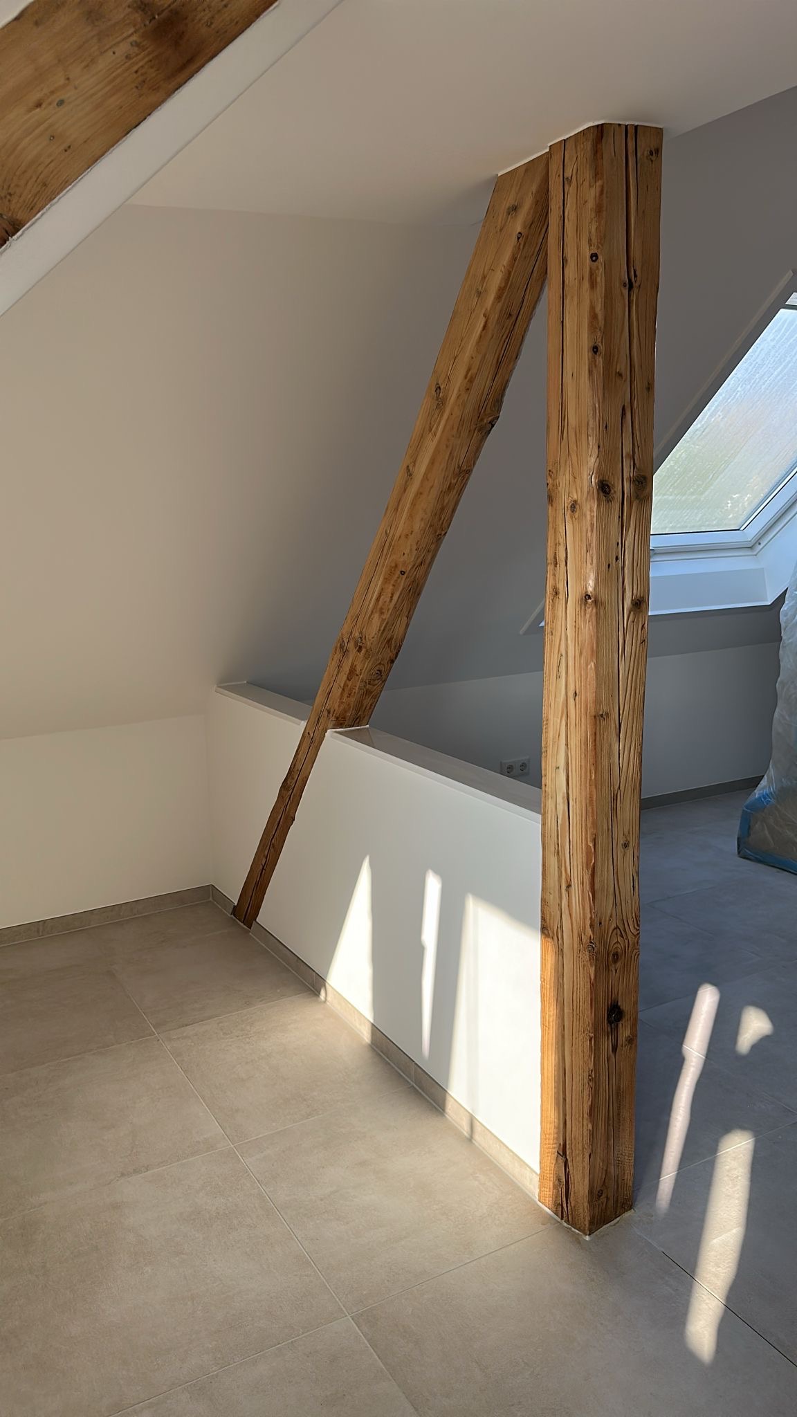 Jasne poddasze po renowacji z drewnianą belką konstrukcyjną, szarą podłogą z płytek i oknem dachowym. Świeżo pomalowane ściany w bieli. Promienie słoneczne podkreślają fakturę drewna.