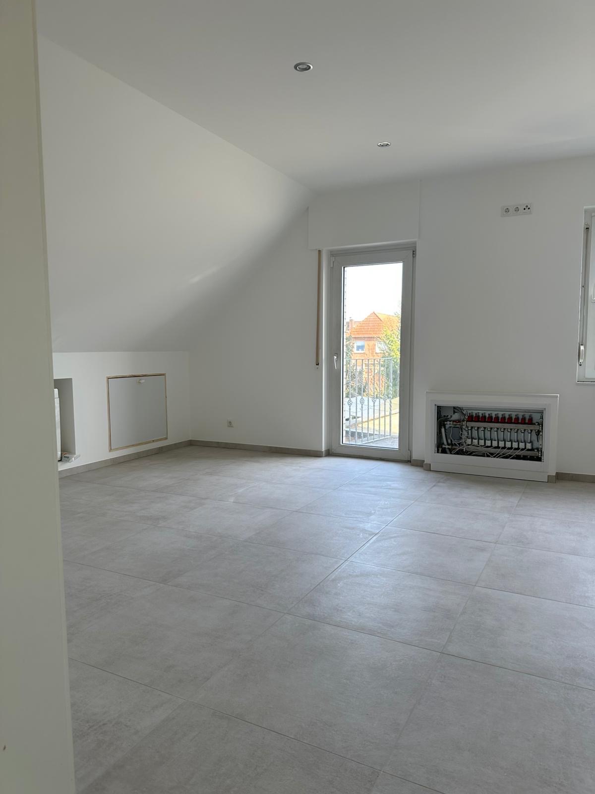 Jasne wnętrze poddasza z szarymi płytkami podłogowymi i białymi ścianami. Widoczna skrzynka z instalacją grzewczą i okno balkonowe z widokiem na dom.