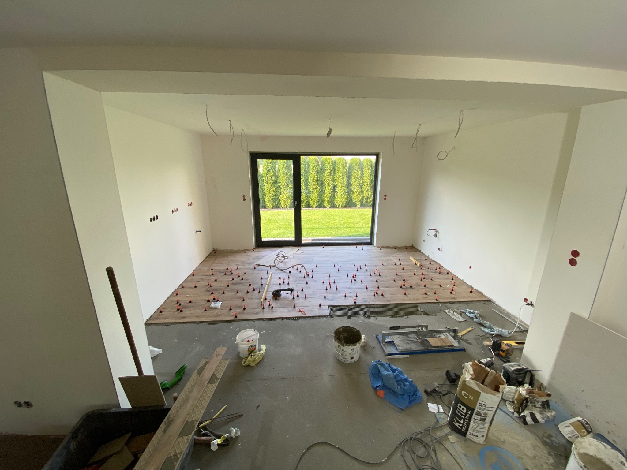 Układanie paneli podłogowych w jasnym wnętrzu z widokiem na zieleń. Widoczne narzędzia i materiały budowlane. Etap prac wykończeniowych.