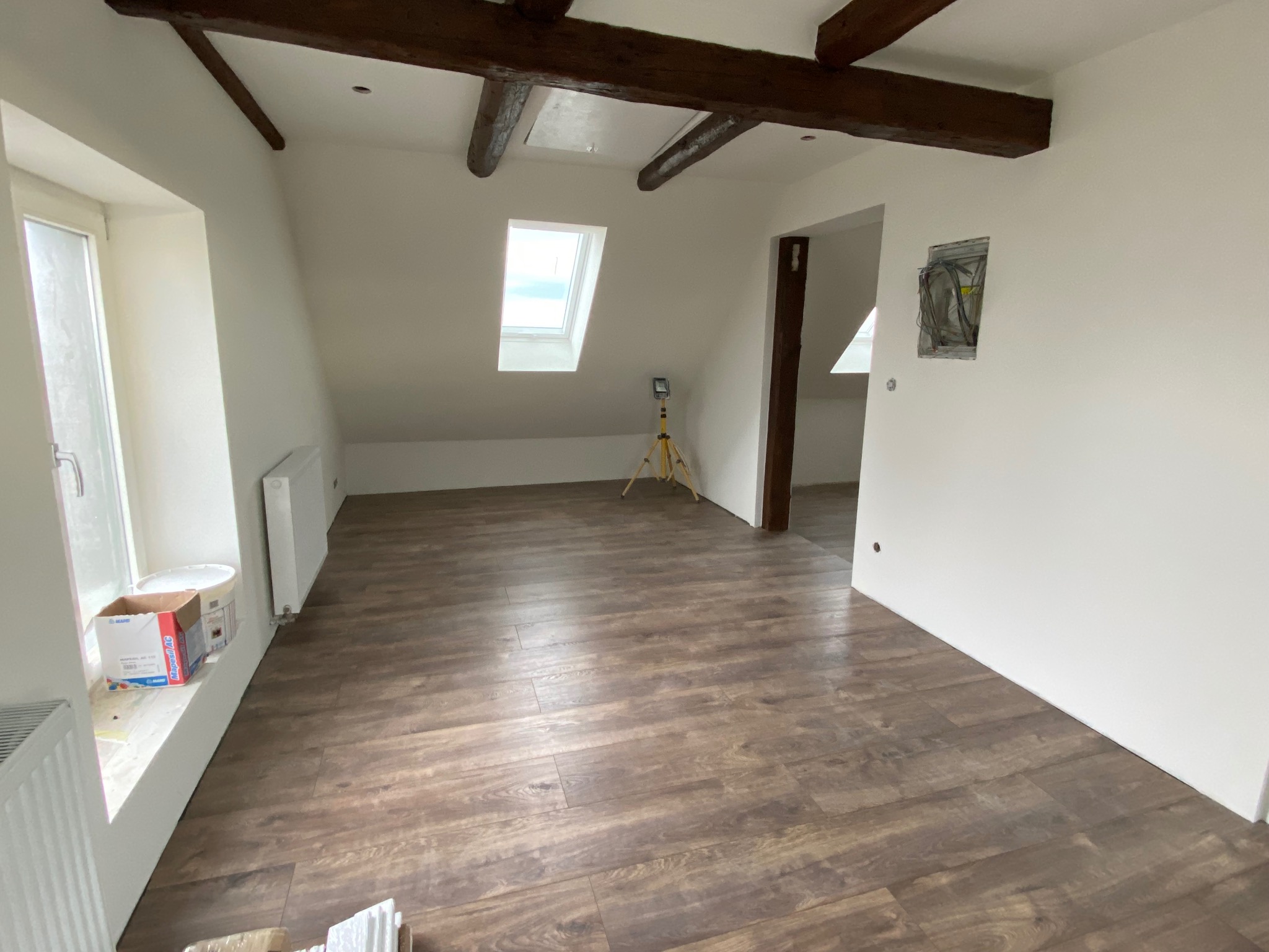 Jasne poddasze po remoncie: drewnopodobne panele podłogowe, białe ściany, widoczne drewniane belki stropowe i okno dachowe. Częściowo widoczne materiały budowlane.