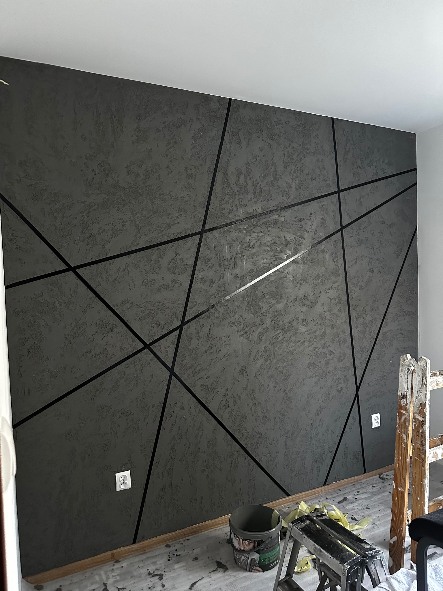 Szara ściana z dekoracyjną fakturą i geometrycznym wzorem z czarnych linii. Widoczne gniazdka elektryczne i drabina malarska. Wykończenie wnętrza z bliska.