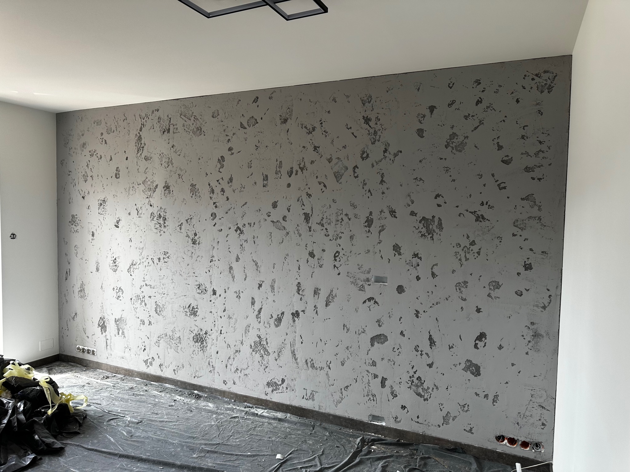 Ściana w pokoju po nałożeniu dekoracyjnej szarej struktury, zabezpieczona folią podłoga, widoczne gniazdka elektryczne i minimalistyczna lampa sufitowa.