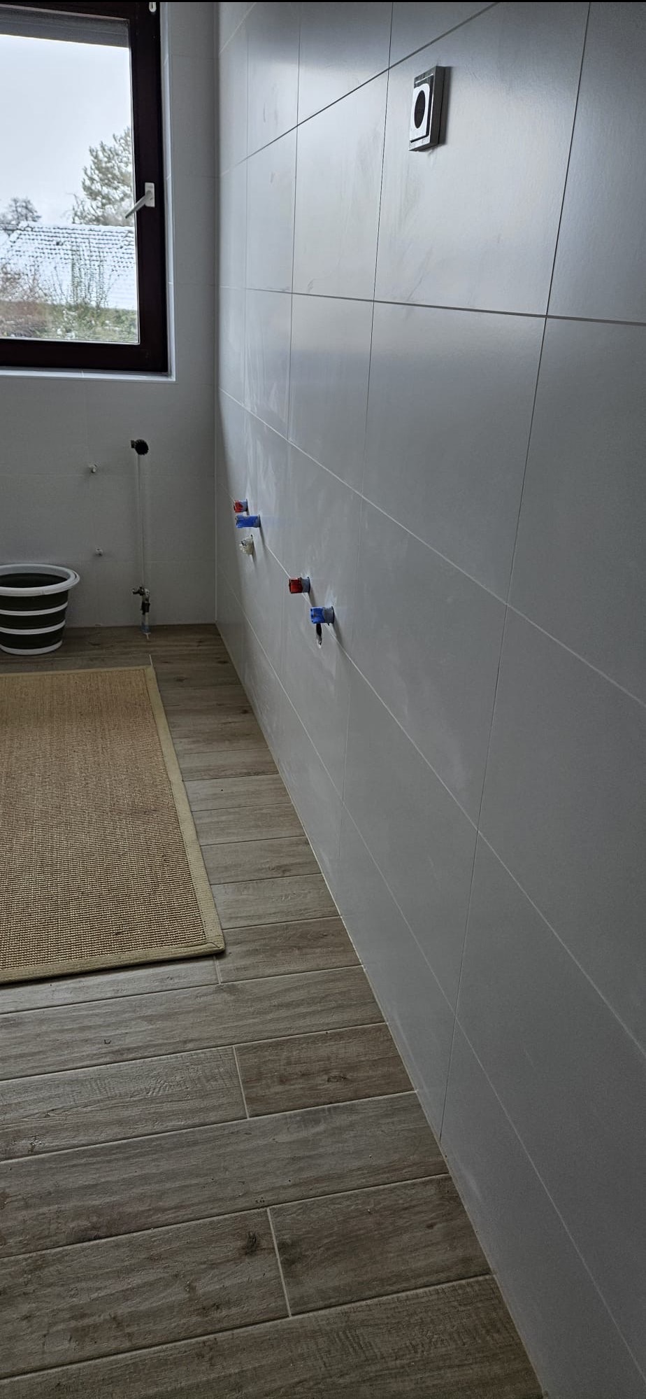 Wykończona łazienka z szarymi płytkami na ścianach i podłodze imitującej drewno. Widoczne przyłącza wodne i okno z widokiem na zewnątrz.