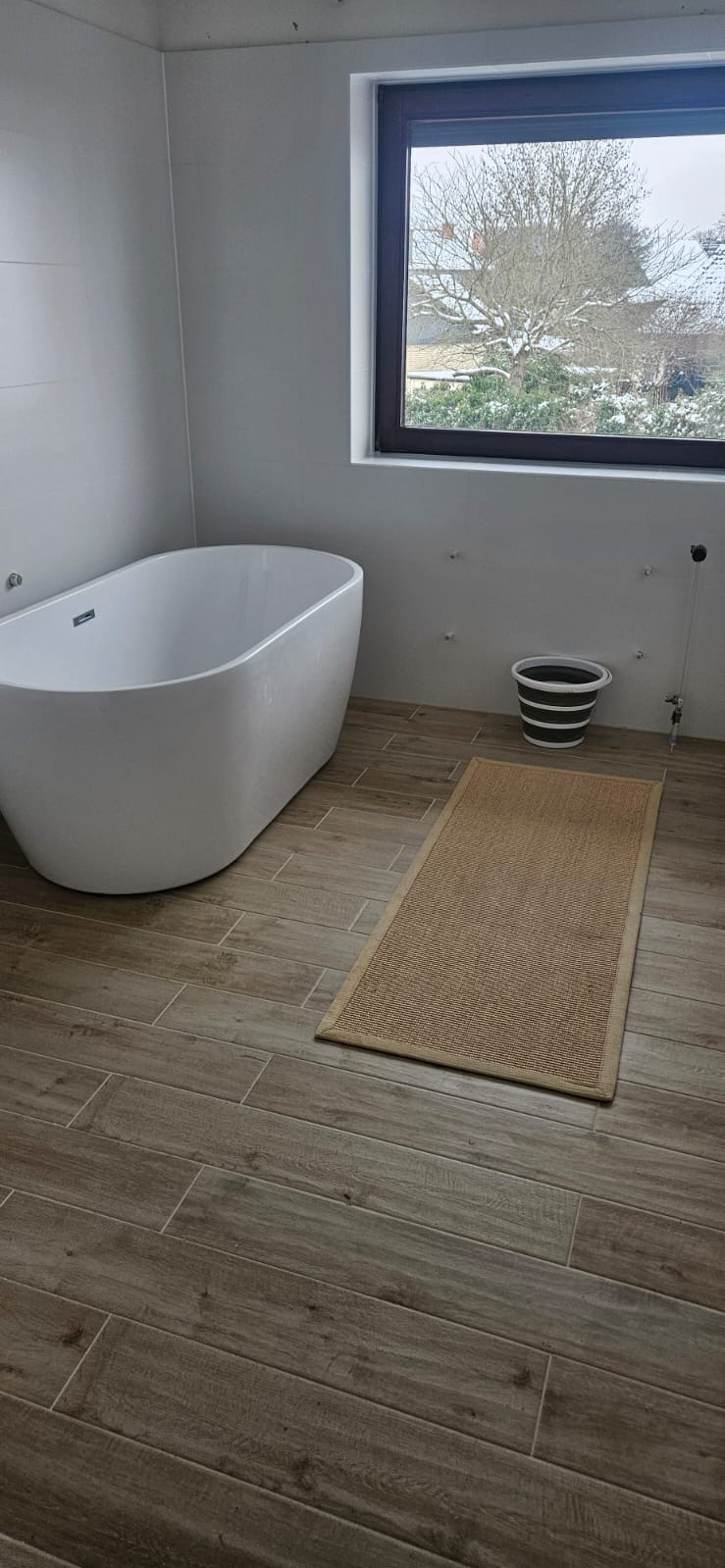 Nowoczesna łazienka z wolnostojącą wanną, drewnopodobnymi płytkami i widokiem na zimowy ogród przez okno. Minimalistyczny design z akcentem naturalnego dywanika.