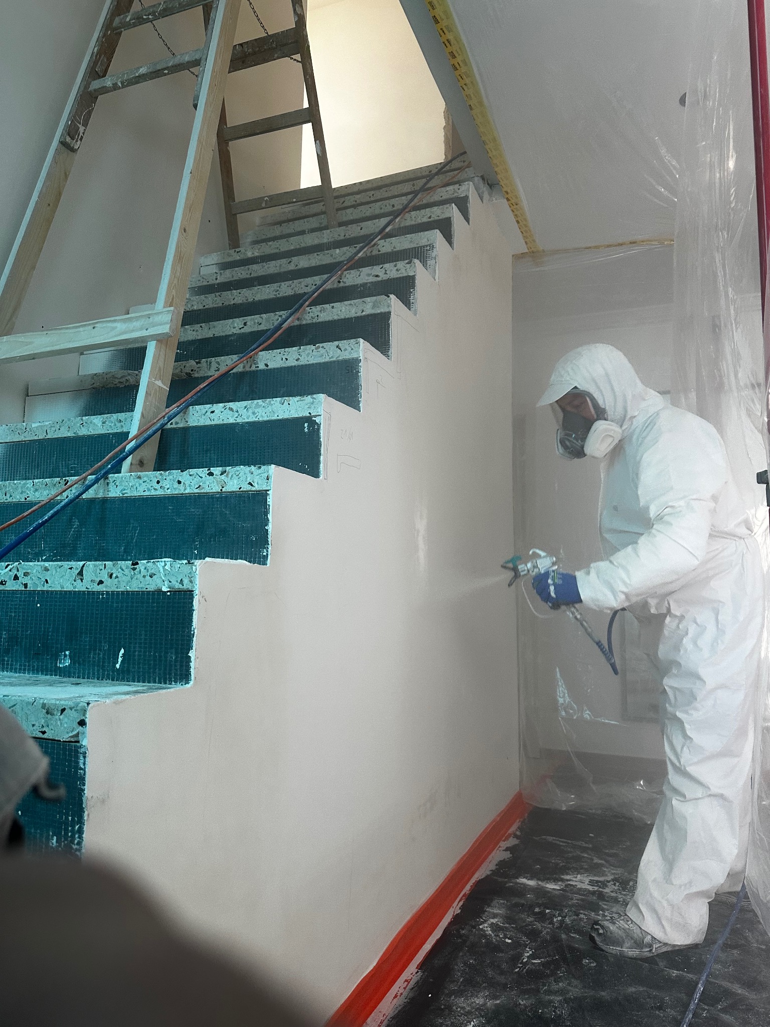 Malowanie natryskowe ściany na klatce schodowej przez pracownika w kombinezonie ochronnym. Widoczna drabina i zabezpieczone schody.