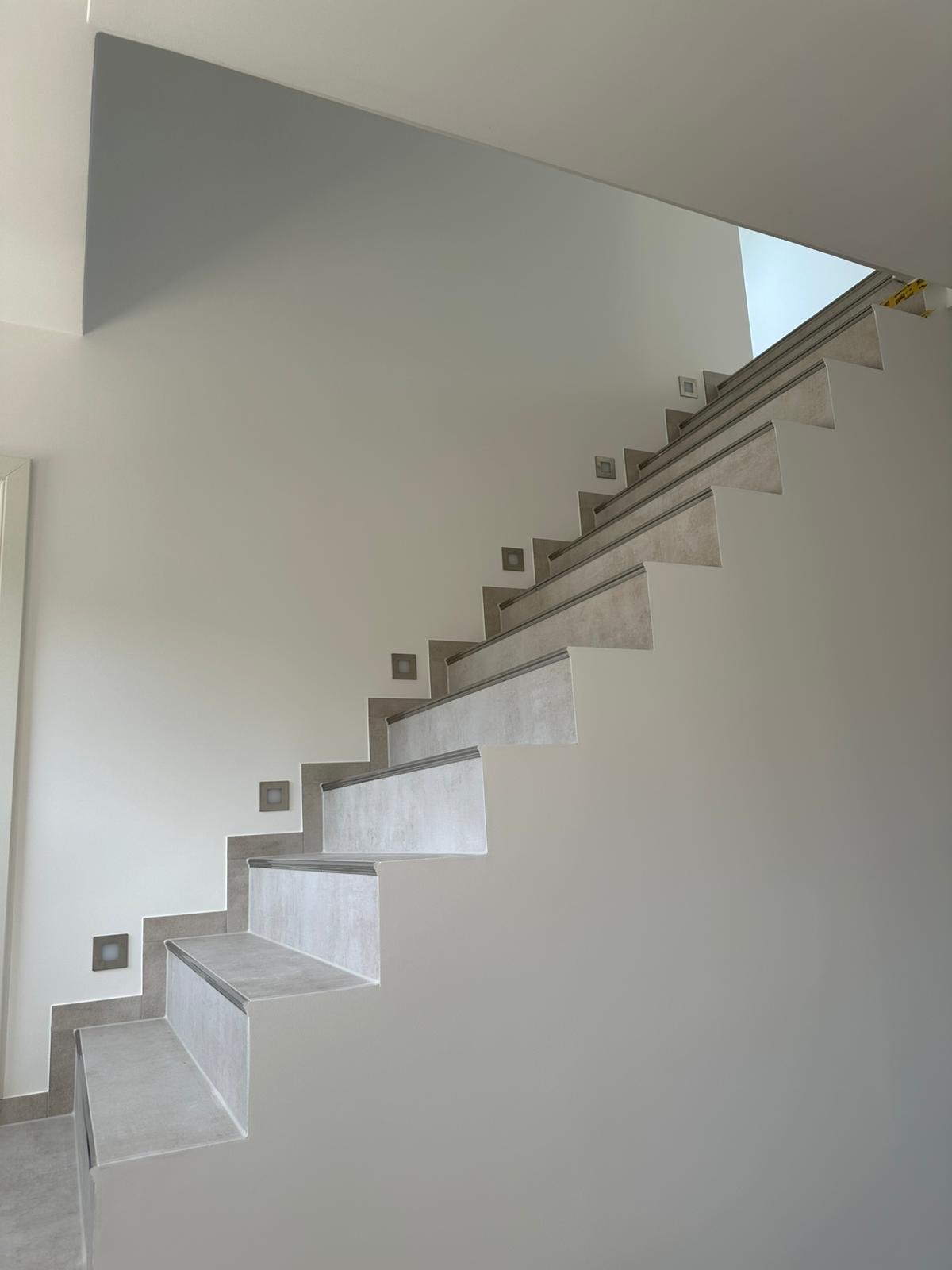 Wykończone schody z szarymi płytkami i oświetleniem punktowym na ścianie. Minimalistyczny design, jasne wnętrze, nowoczesne wykończenie.