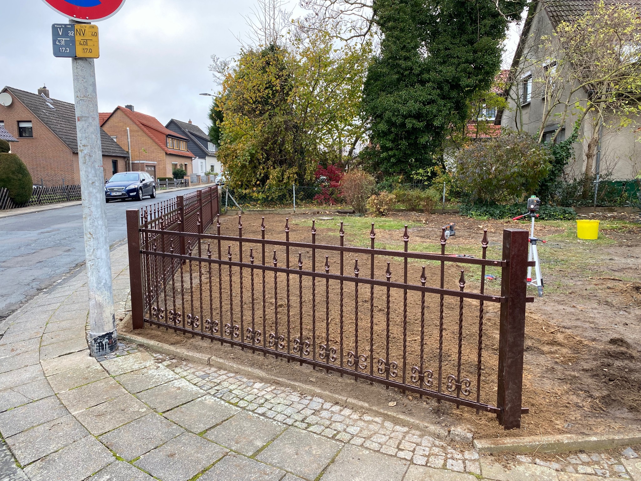 Montaż ozdobnego, brązowego ogrodzenia metalowego na posesji. Widoczny fragment chodnika, ulica z domami w tle i słup z tablicami informacyjnymi.