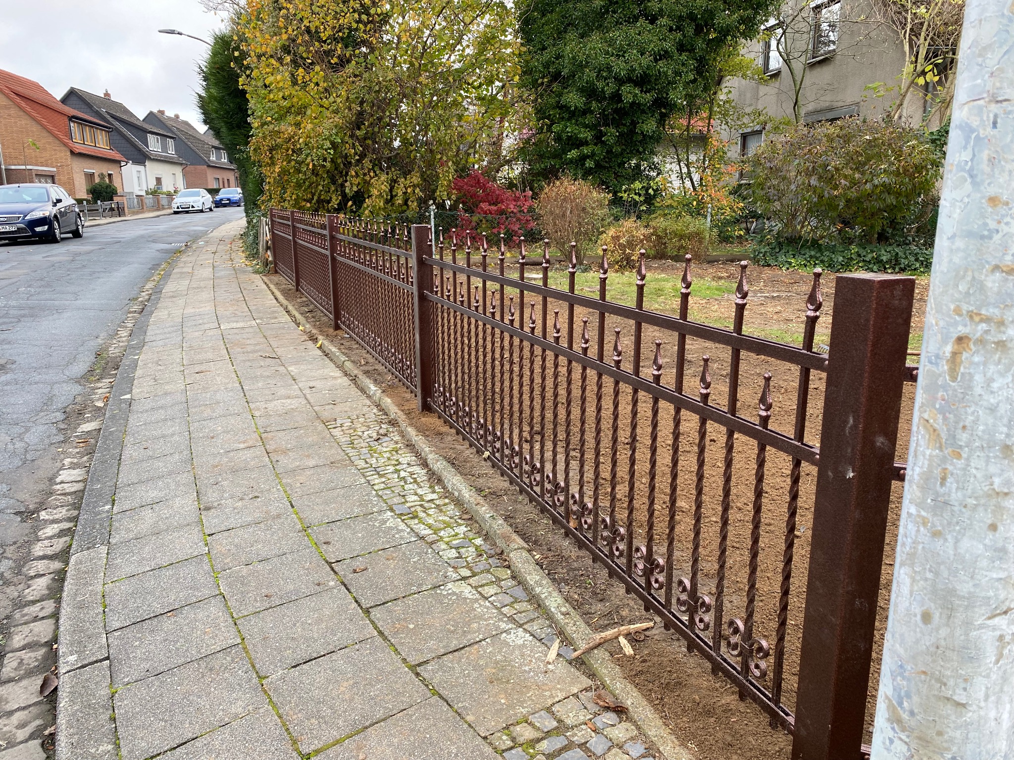 Nowoczesne, brązowe ogrodzenie metalowe z ozdobnymi detalami, oddzielające chodnik od posesji z zielenią. Widok na ulicę z domami w tle.