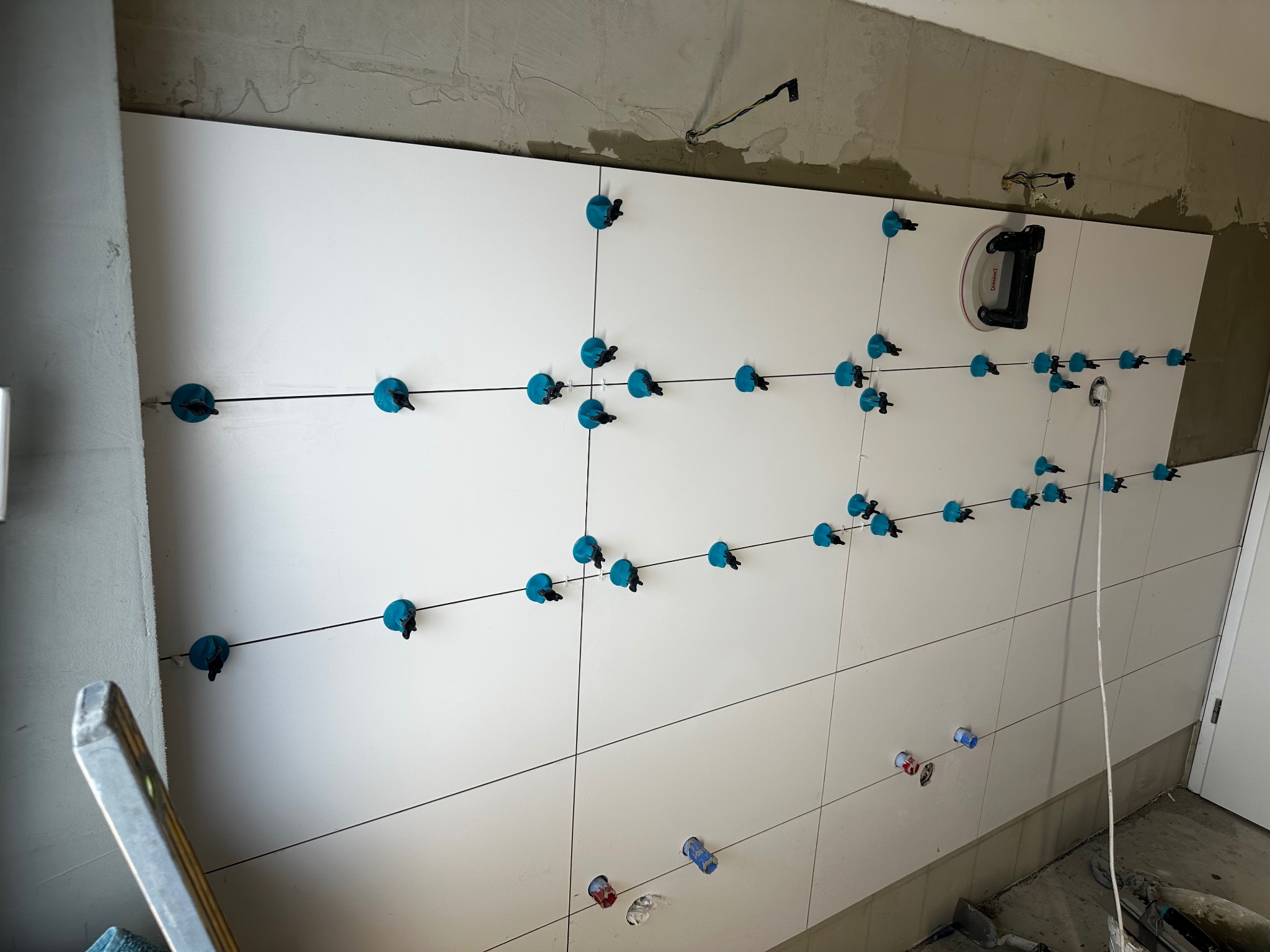 Białe płytki ścienne w trakcie montażu, z użyciem systemu poziomowania płytek (niebieskie klipsy i kliny). Widoczne otwory na instalacje. Ściana częściowo wykończona.