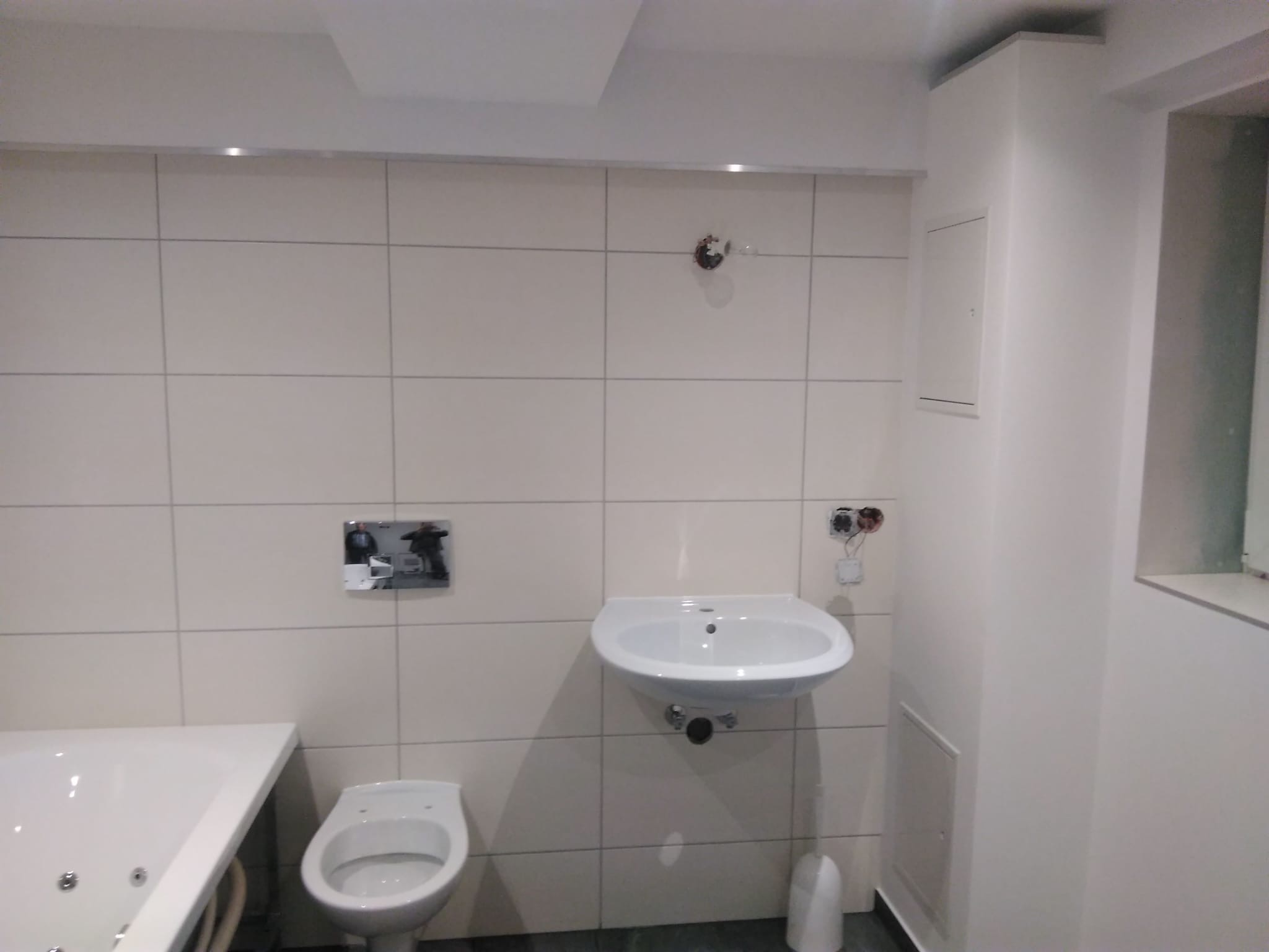 Jasna łazienka po remoncie: białe płytki, umywalka, toaleta i wanna z hydromasażem. Widoczne niedokończone podłączenia elektryczne.