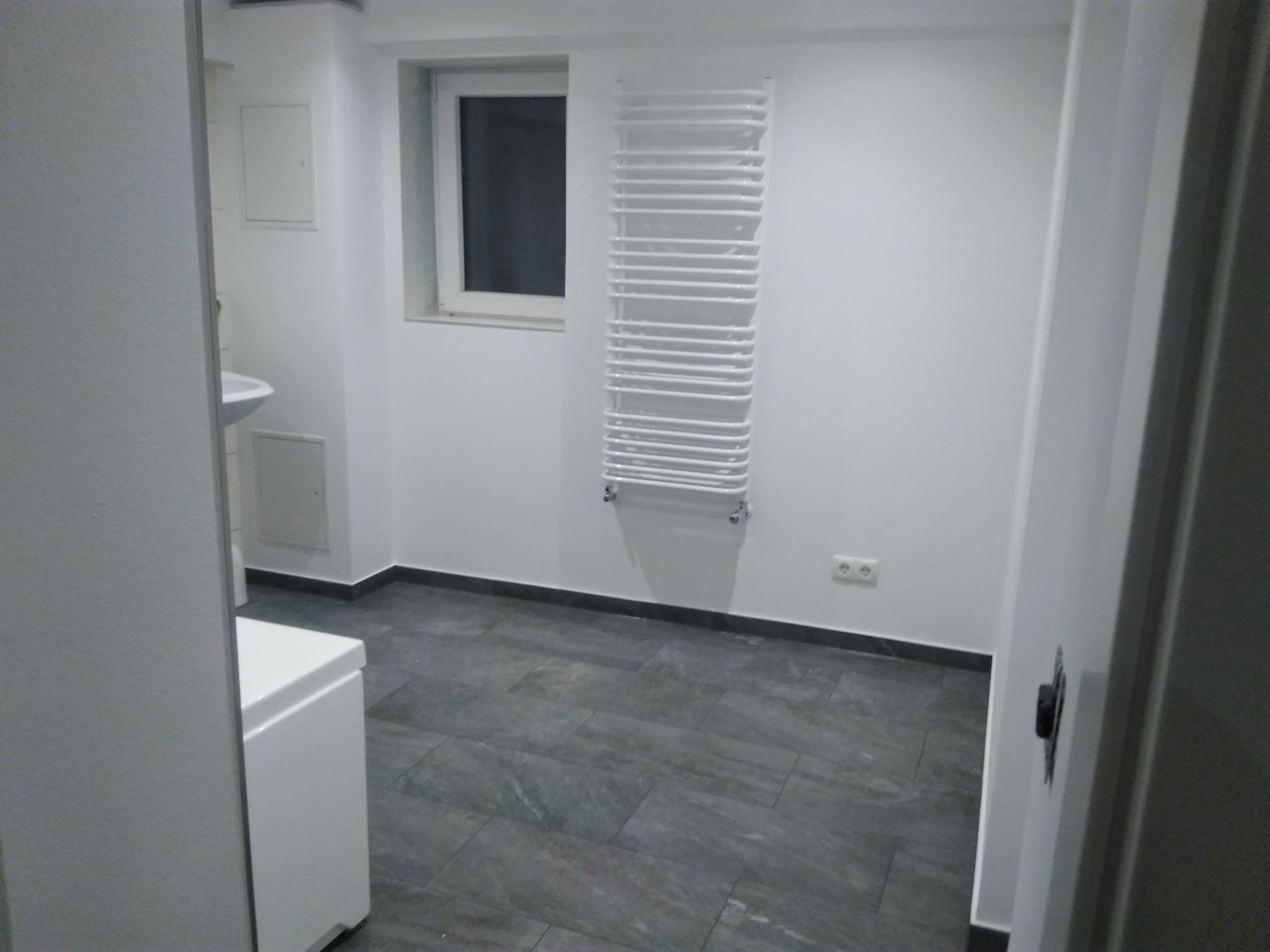 Wykończona łazienka w odcieniach bieli i szarości. Na ścianie grzejnik drabinkowy, podłoga wyłożona płytkami imitującymi kamień. Widok zza uchylonych drzwi.