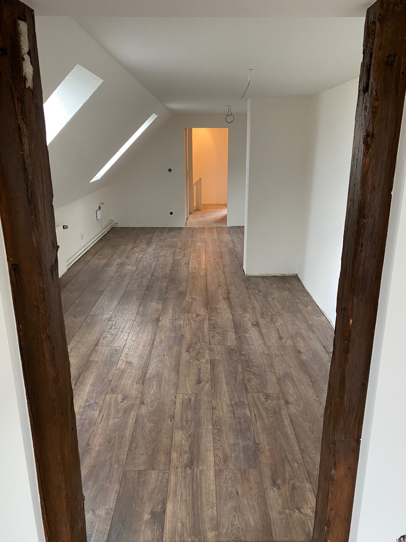 Jasne poddasze z nową, brązową podłogą z paneli, świeżo pomalowanymi ścianami i drewnianą belką po prawej stronie kadru. Widok na kolejne pomieszczenie.