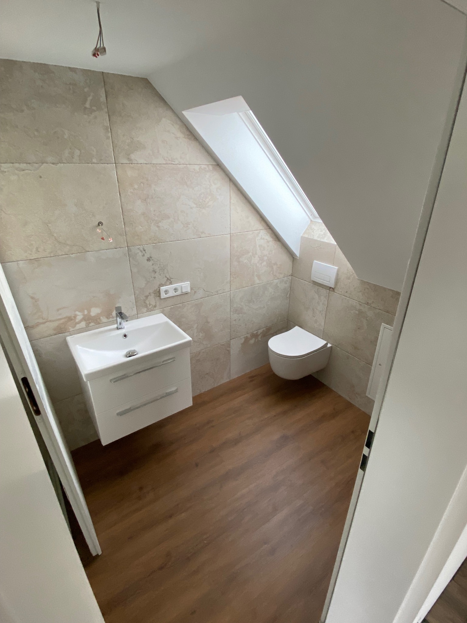 Wykończona łazienka na poddaszu z beżowymi płytkami imitującymi kamień, brązową podłogą, białą umywalką z szafką i podwieszaną toaletą. Okno dachowe w skosie.