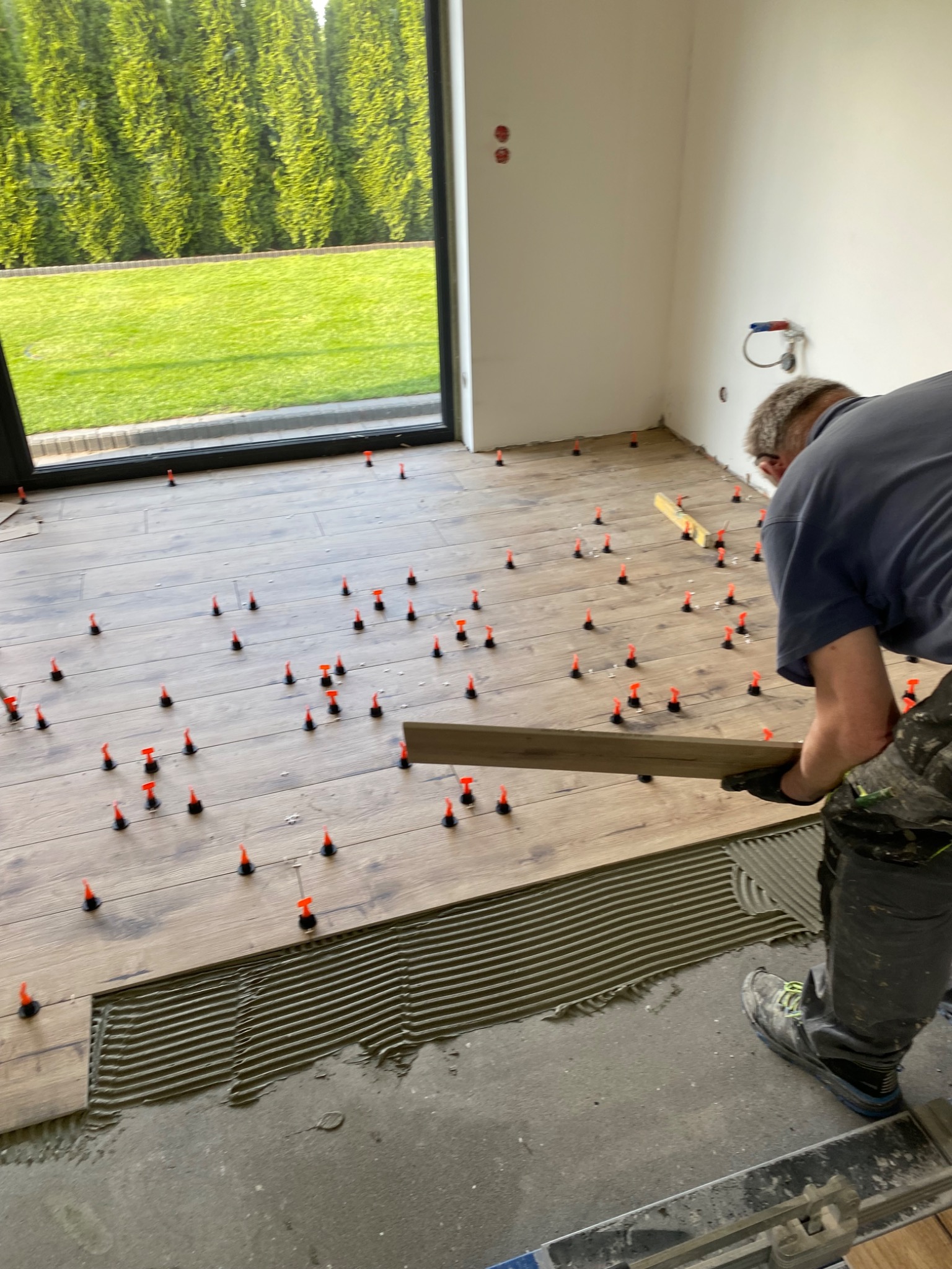 Układanie paneli podłogowych z użyciem poziomicy i klipsów dystansowych w jasnym wnętrzu z dużym oknem na ogród. Widoczny fragment nałożonego kleju.