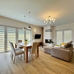 NKK Nieruchomości Natalia Kucharska - Jasne wnętrze salonu z jadalnią: drewniany stół z krzesłami, szara sofa, kominek i duże okna z żaluzjami. Na podłodze płytki imitujące drewno. Widoczny zegar na ścianie.