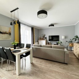 NKK Nieruchomości Natalia Kucharska - Jasne wnętrze salonu z jadalnią: stół z kwiatami, szare ściany, drewniana podłoga, sofa i obrazy. Nowoczesny design z akcentami żółci.