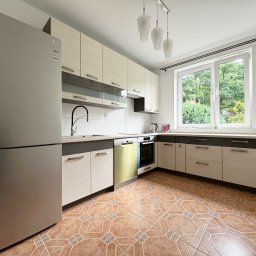 NKK Nieruchomości Natalia Kucharska - Jasna kuchnia z beżowymi szafkami, brązową podłogą i dużym oknem z widokiem na zieleń. Lodówka LG po lewej. Uwagę przyciągają wzorzyste płytki podłogowe.