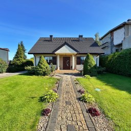 NKK Nieruchomości Natalia Kucharska - Ujęcie domu jednorodzinnego z ogrodem, z kamienną ścieżką prowadzącą do wejścia. Widoczne elementy architektury i zieleni w słoneczny dzień.