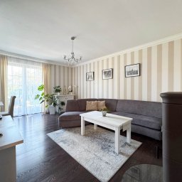 NKK Nieruchomości Natalia Kucharska - Jasny salon z szarą sofą narożną, białym stolikiem kawowym i wzorzystym dywanem. Na ścianie w paski wiszą trzy czarno-białe zdjęcia. Widoczne rośliny doniczkowe i komoda.