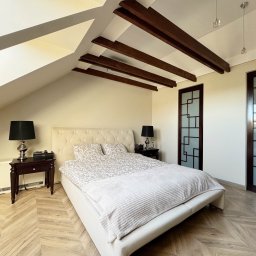 NKK Nieruchomości Natalia Kucharska - Jasna sypialnia na poddaszu z drewnianymi belkami stropowymi, parkietem w jodełkę i łóżkiem z beżowym zagłówkiem. Widoczne drzwi przesuwne z geometrycznym wzorem.