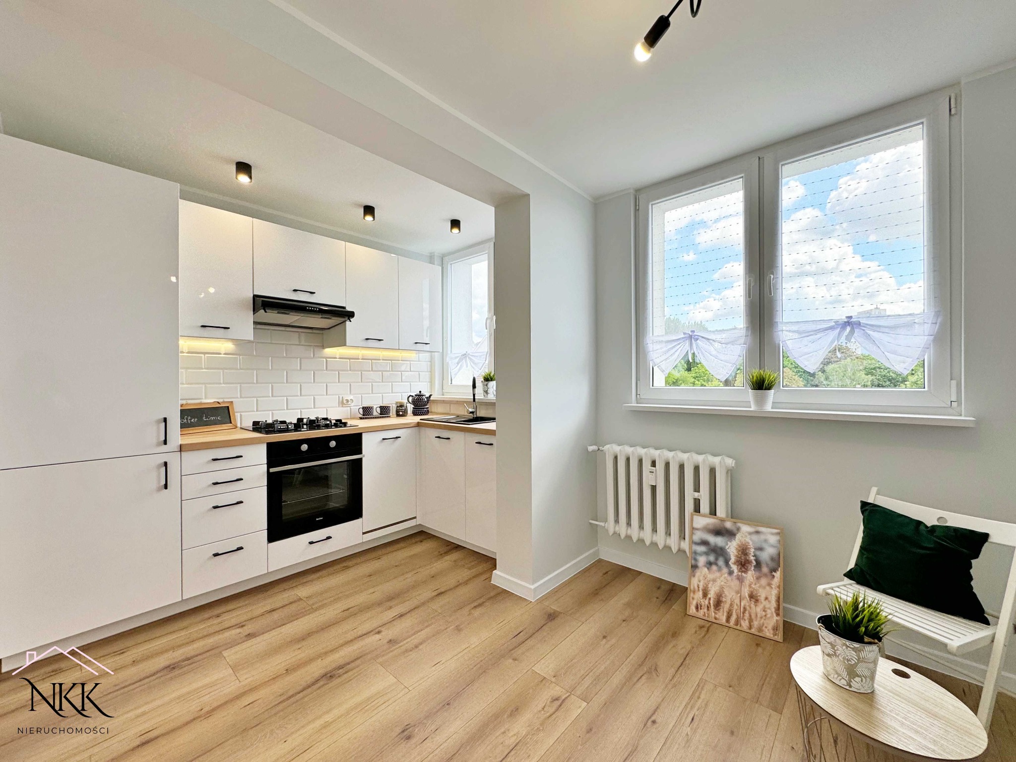 Jasna kuchnia z białymi szafkami, drewnianym blatem i podłogą. W tle okno z firankami i dekoracyjny grzejnik. W rogu krzesło z poduszką i stolik z rośliną.