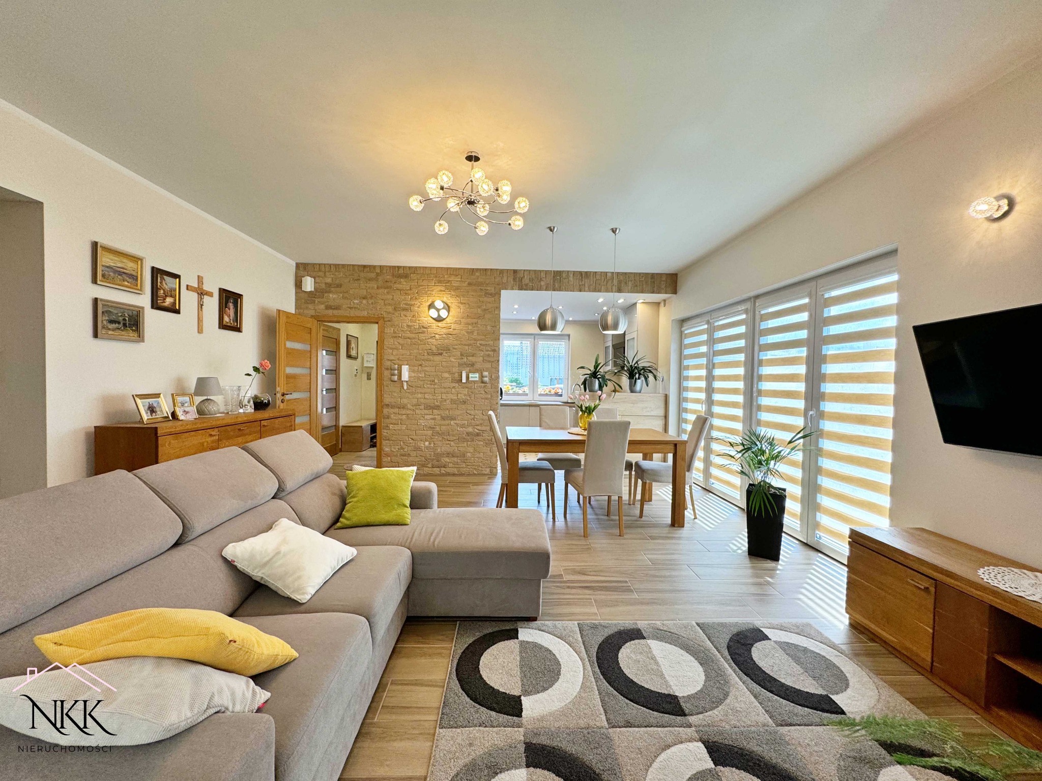 Jasne wnętrze domu z szarą sofą, drewnianym stołem i ceglaną ścianą. Widoczne dekoracje, dywan w geometryczne wzory i roślina doniczkowa przy oknie.