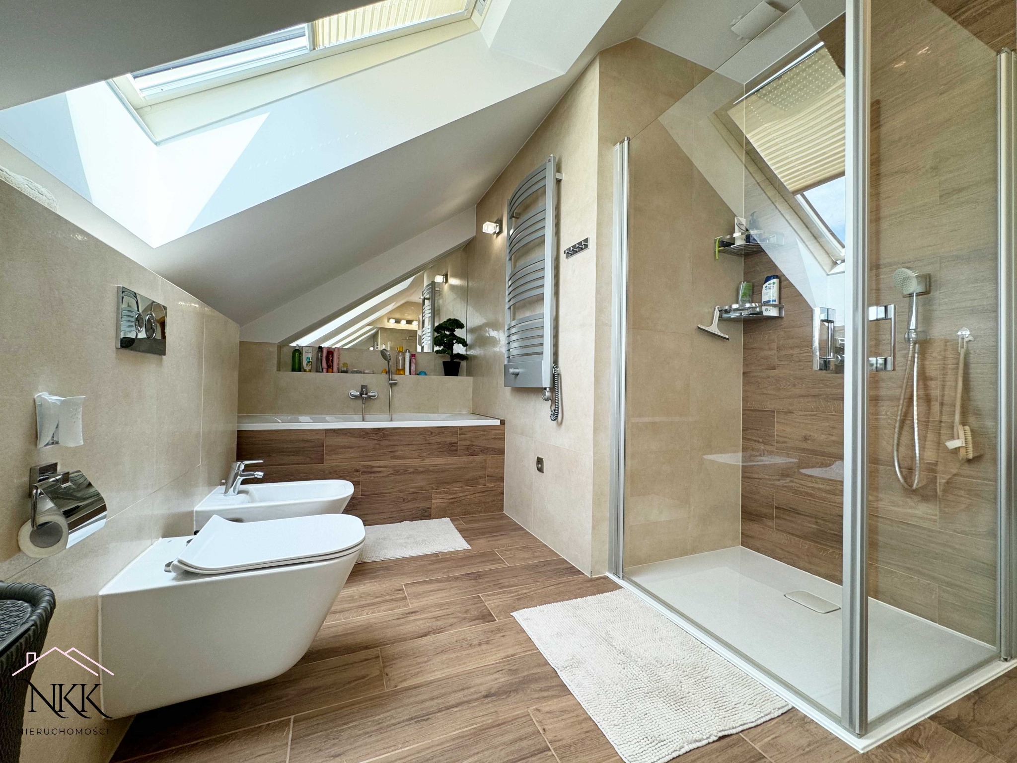 Jasna łazienka na poddaszu z kabiną prysznicową, bidetem i oknem dachowym. Beżowe płytki imitujące drewno, minimalistyczny design. Widok z perspektywy wejścia.
