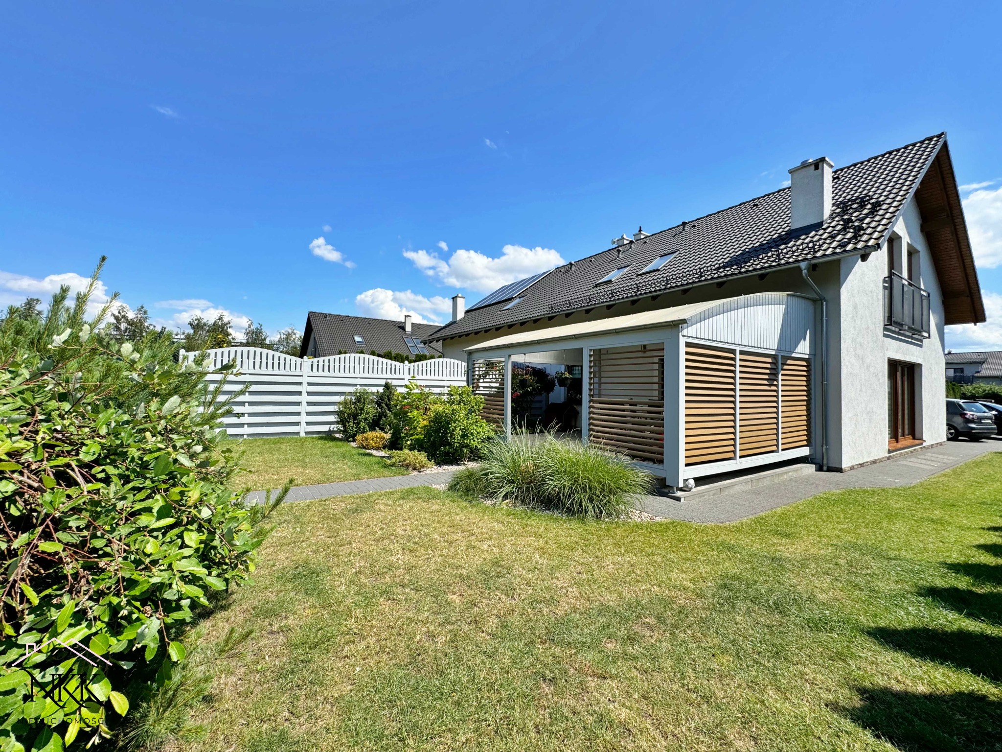 Nowoczesny dom jednorodzinny z ogrodem w słoneczny dzień. Widoczne panele słoneczne na dachu, drewniane żaluzje na tarasie i białe ogrodzenie.