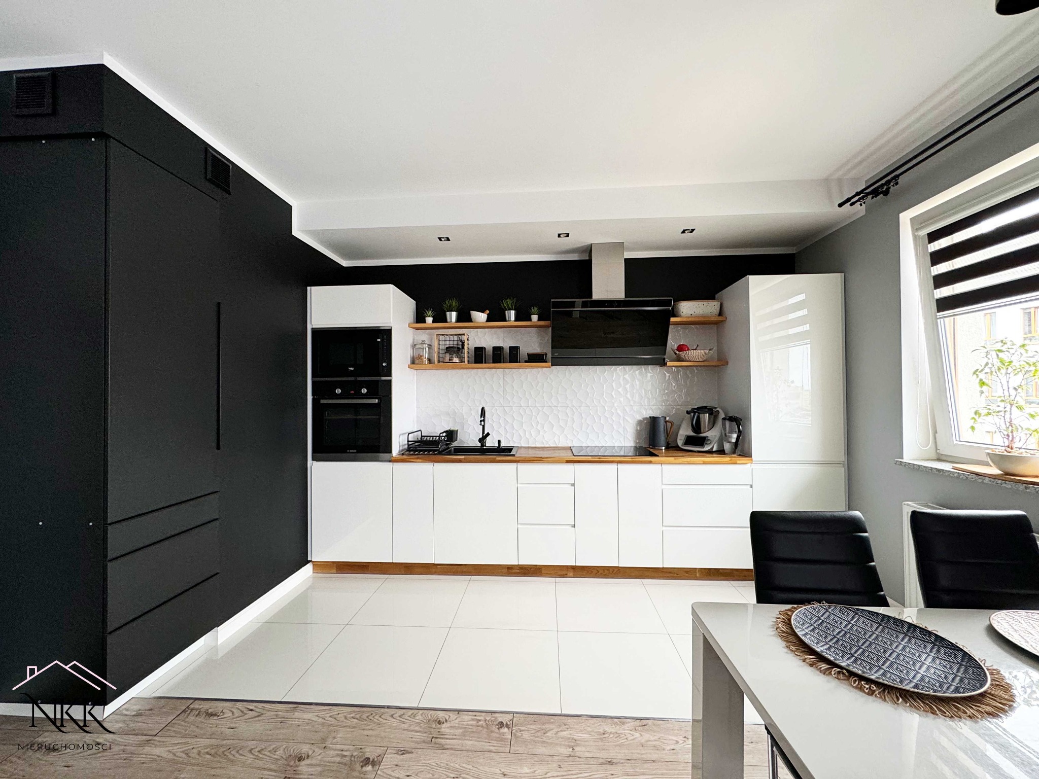 Nowoczesna kuchnia w bieli i czerni z drewnianymi akcentami. Białe płytki na podłodze i ścianie, czarne szafki i drewniane półki. Minimalistyczny design z roślinami.