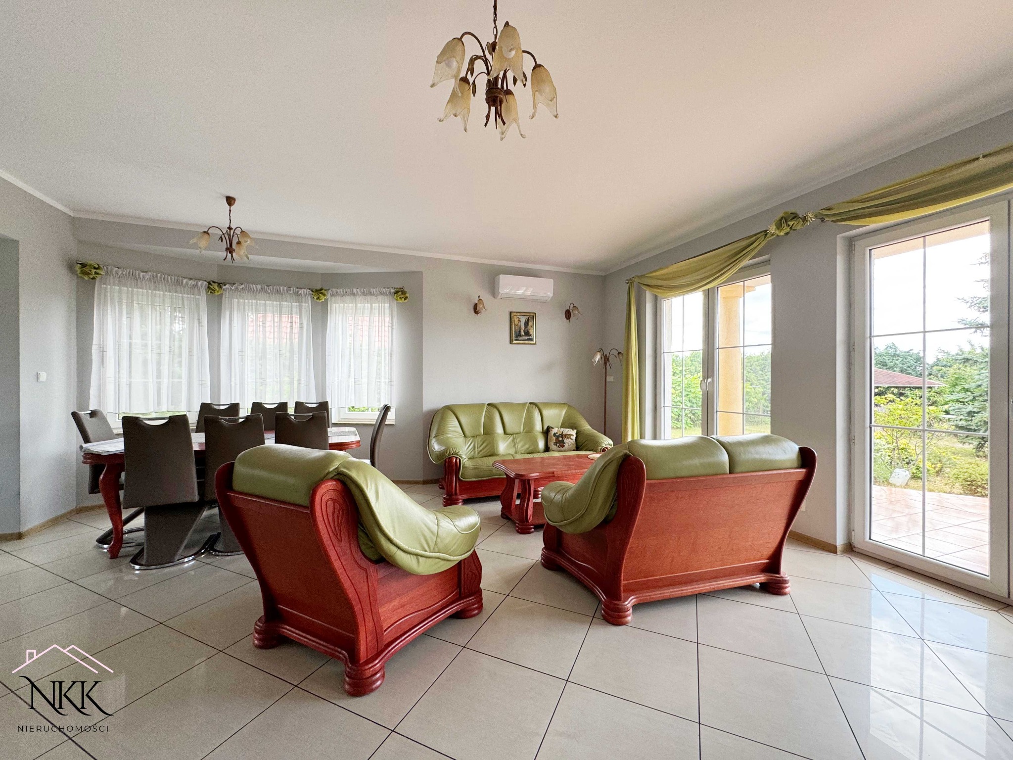 Jasne wnętrze domu z zieloną skórzaną sofą i fotelami, drewnianym stołem jadalnym i dużymi oknami z dekoracyjnymi zasłonami. Widok na ogród.