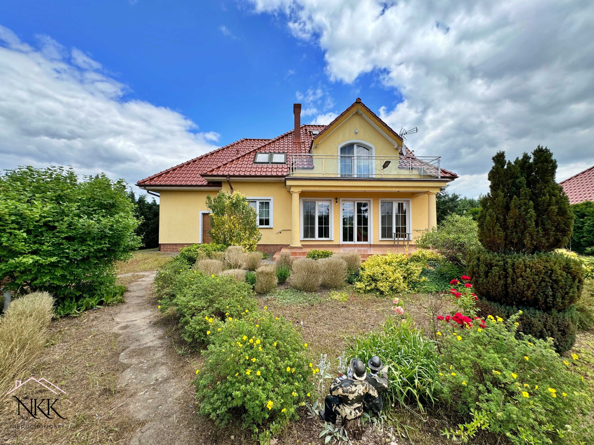 Żółty dom jednorodzinny z czerwonym dachem i balkonem, otoczony zielenią i kwiatami. Na pierwszym planie dekoracja ogrodowa w postaci figurek.