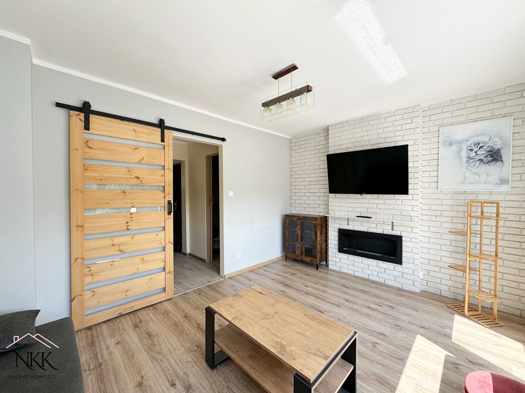 Jasne wnętrze salonu z drewnianymi drzwiami przesuwnymi, ceglaną ścianą z kominkiem i telewizorem. Widoczny stolik kawowy i drewniana półka. Nowoczesny design.