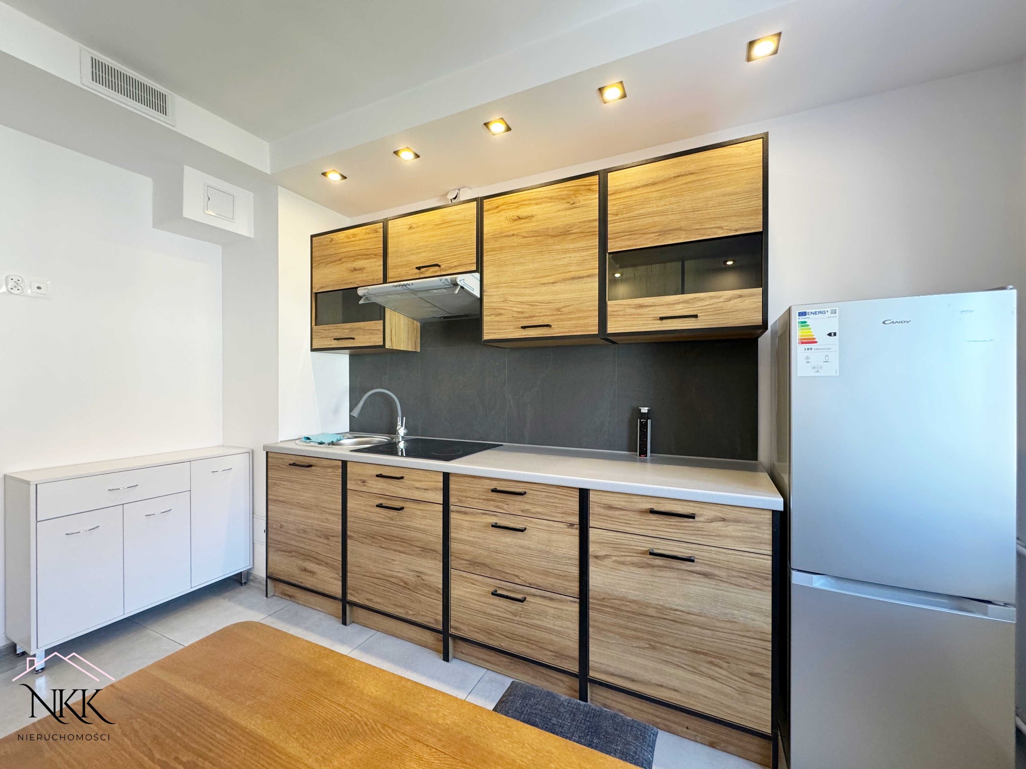 Nowoczesna kuchnia z drewnianymi szafkami i czarnymi detalami, oświetlona punktowo, z widoczną lodówką i fragmentem stołu. Minimalistyczny design.