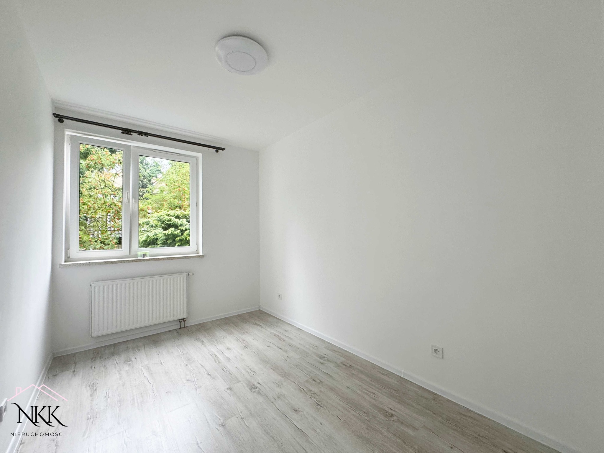 Jasne, puste wnętrze pokoju z oknem wychodzącym na zieleń. Parkiet w odcieniach szarości, białe ściany i grzejnik pod oknem. Minimalistyczny design, widoczny kontakt elektryczny.