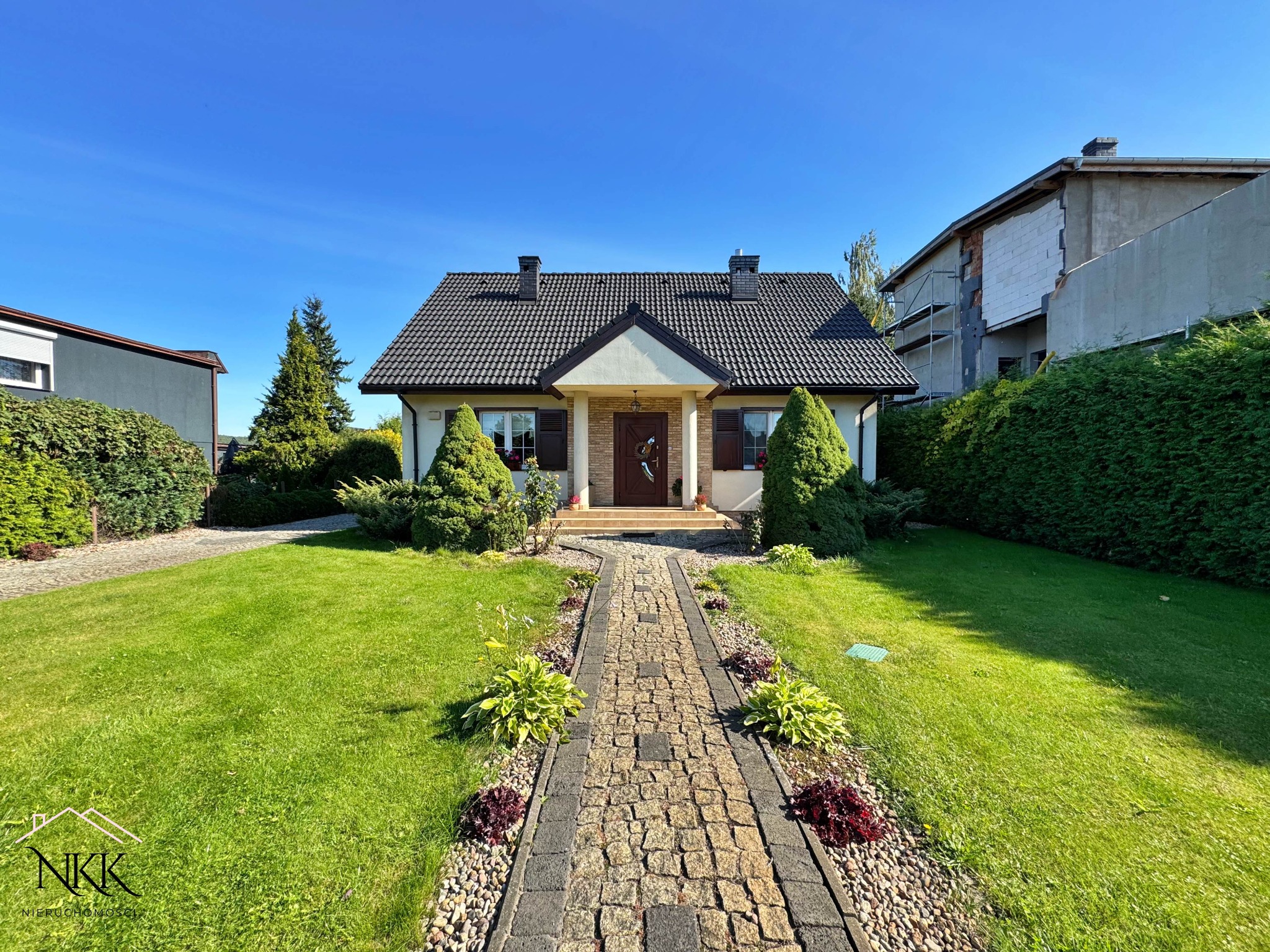 Ujęcie domu jednorodzinnego z ogrodem, z kamienną ścieżką prowadzącą do wejścia. Widoczne elementy architektury i zieleni w słoneczny dzień.