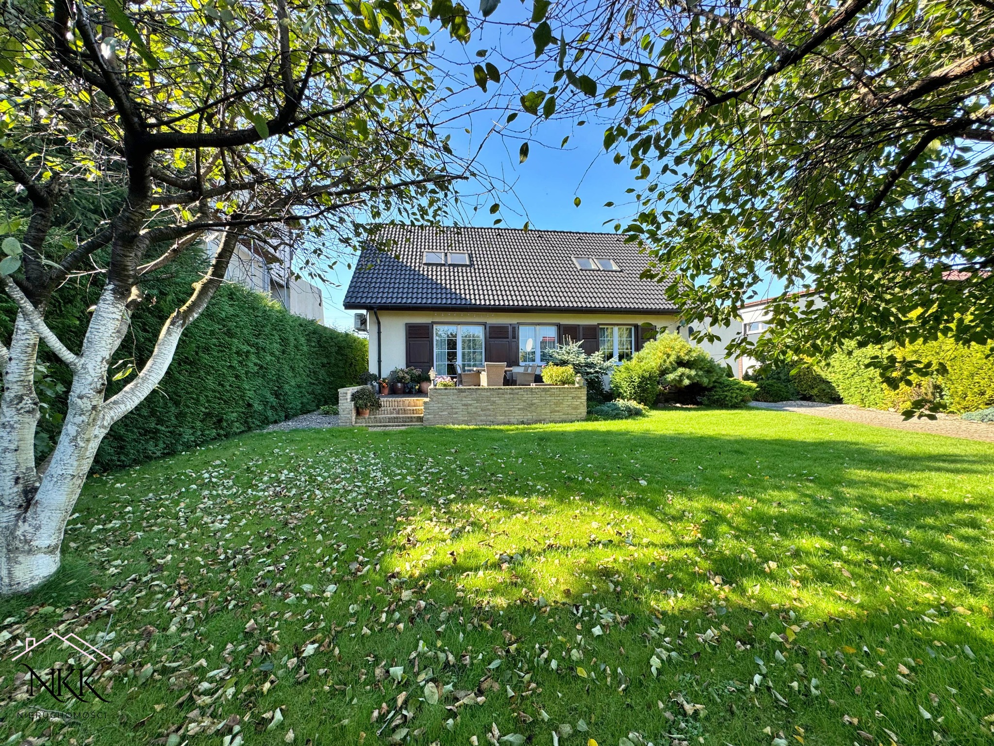 Dom jednorodzinny z brązowym dachem i oknami, otoczony zielonym trawnikiem z opadłymi liśćmi, widziany zza drzew w słoneczny dzień.