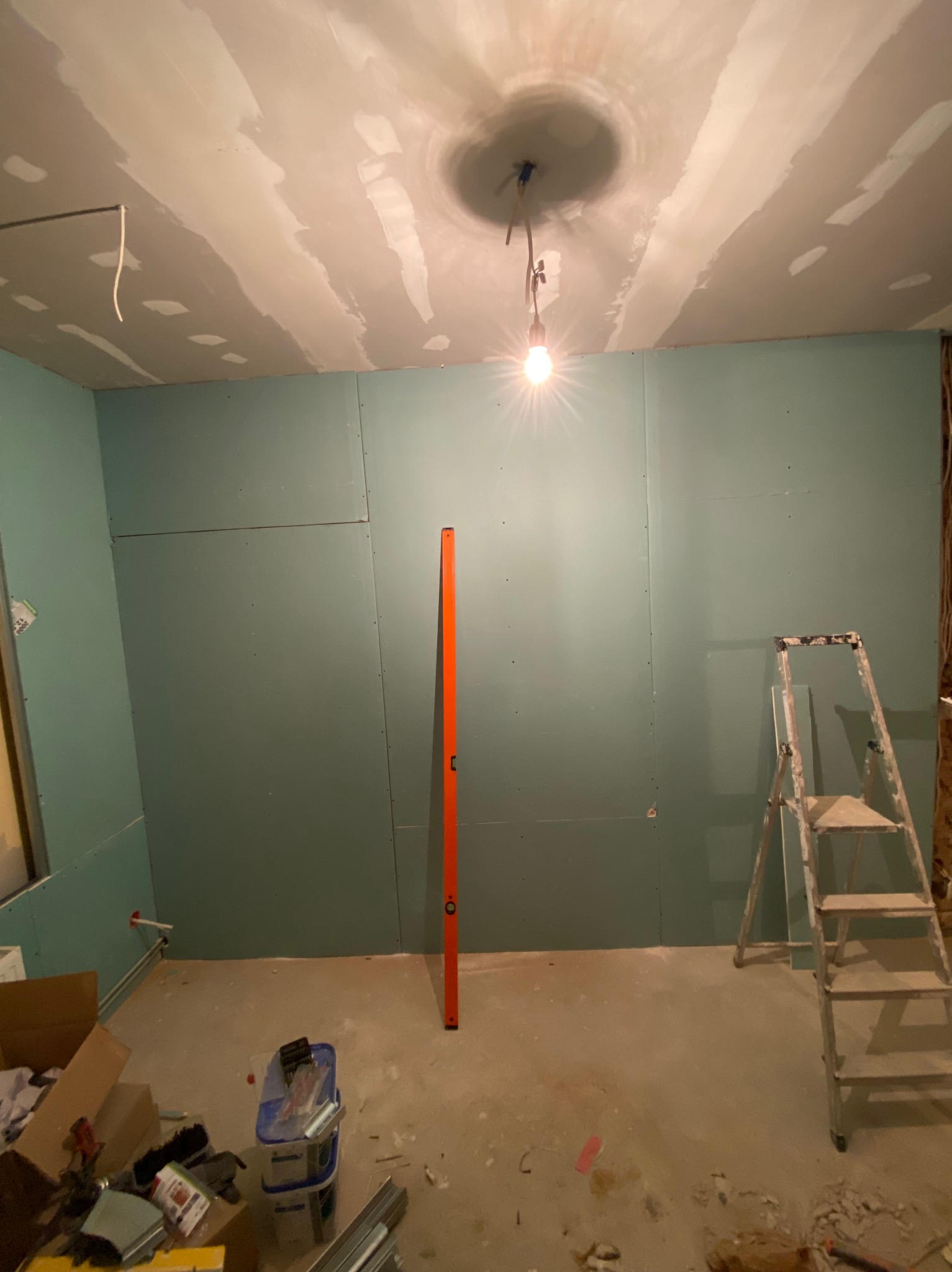 Pokój w trakcie montażu płyt karton-gips na ścianach i suficie. Widoczna drabina, poziomica i narzędzia. Sufit z widocznymi łączeniami płyt i lampą.