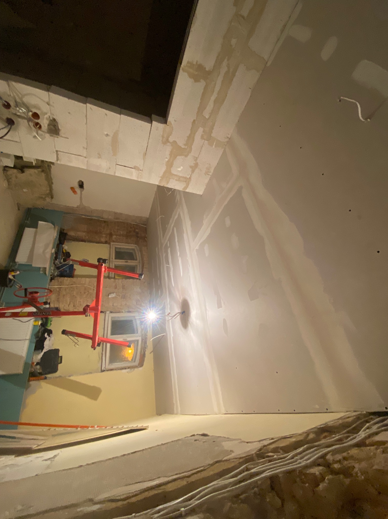 Wnętrze w trakcie prac: ściany z płyt gipsowo-kartonowych, widoczne łączenia szpachlowane, podnośnik do płyt. Prace wykończeniowe w pomieszczeniu z oknami.