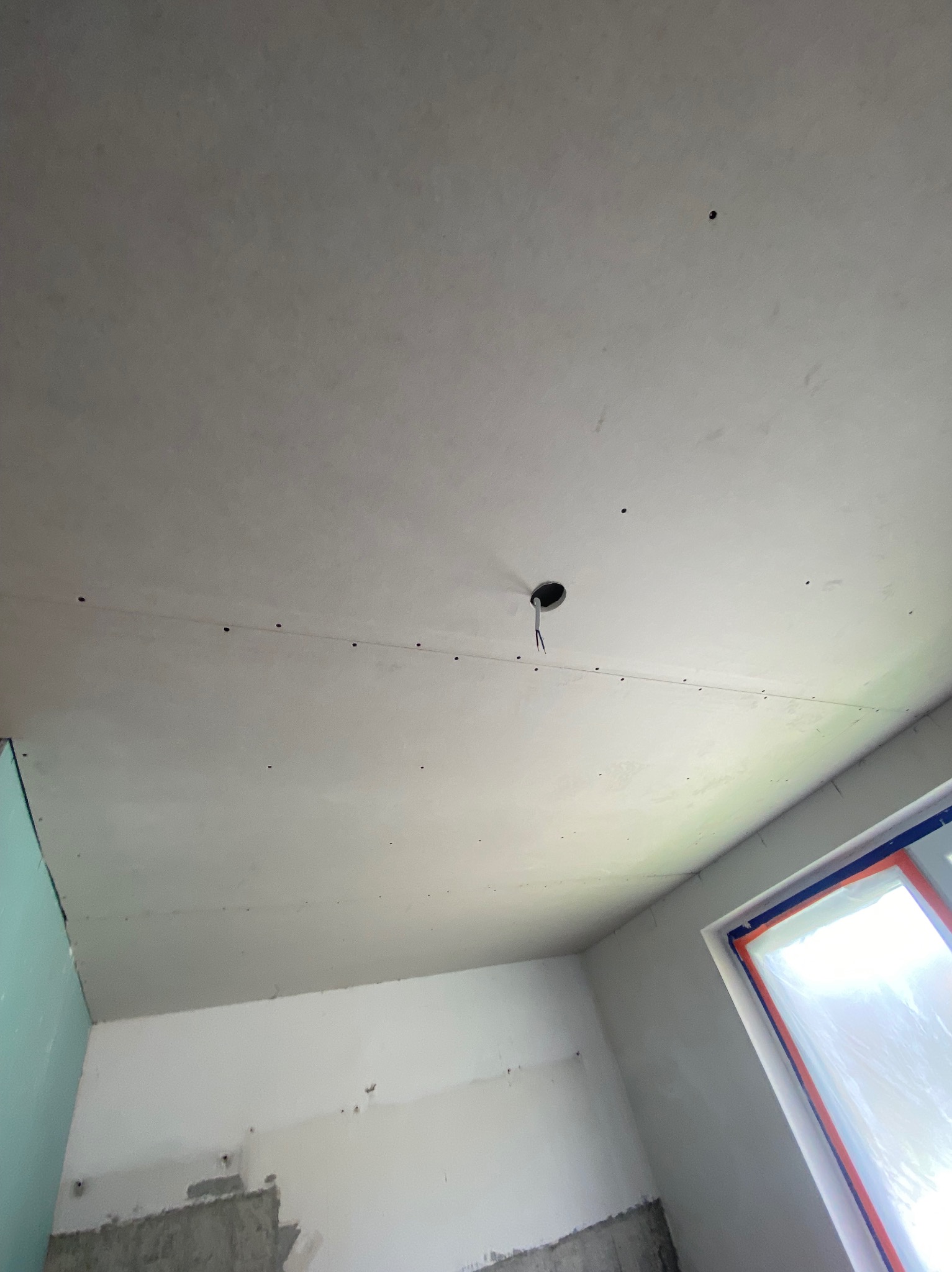 Sufit z płyt kartonowo-gipsowych z otworem na oświetlenie, widoczne łączenia płyt i ślady szpachlowania, okno zabezpieczone folią malarską.